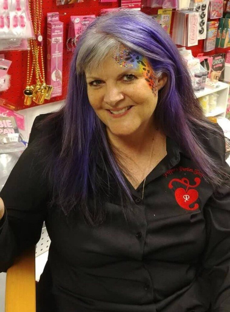 Kathi Pepper smiling in the Pepper's Parties boutique with purple hair and bright face art
